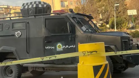 Supplied An armoured vehicle is parked in front of yellow tape and a yellow plastic street blockade.
