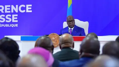 DR Congo Presidency/X President Félix Tshisekedi, dressed in a blue suit, sits in a white chair behind a lectern as he addresses a press conference, with the national flag visible behind him and members of the audience facing the podium