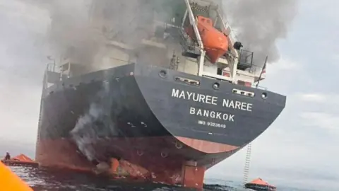 Royal Thai Navy Smoke rises above a cargo ship with 'Mayuree Naree Bangkok' written on it on 11 March 2026. Rafts are floating in the sea around it.