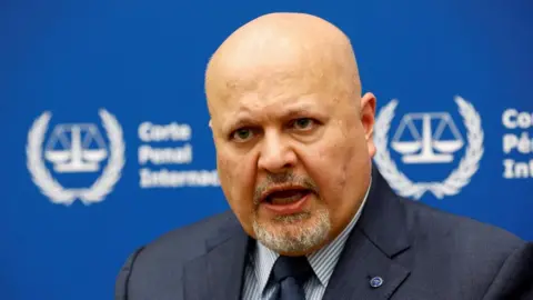 Reuters International Criminal Court Prosecutor Karim Khan speaks during an interview while sitting in front of a blue sign with blurry white writing and the symbol of the ICC - the scales of justice surrounded by a laurel wreath.