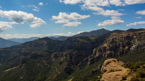 Getty Images An image depicting the hills of Tremp in Catalonia. Large green and brown mountains stand in front of a blue sky with several clouds.