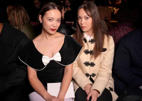 Getty Images Laufey and her sister Junia sit side by side during a public event. Laufey is dressed in an elegant black gown with a white bow on the neckline; while Junia is in a tan button-up jacket, with ornate black clasps