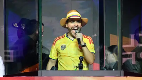 Getty Images Abelardo de la Espriella speaks behind bulletproof glass during a campaign rally in Medellin. He wears a Colombia football shirt and striped hat.