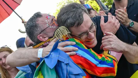 EPA/Shutterstock A person wearing a jacket in the colours of the pride flag hugs Budapest Mayor Gergely Karacsony, who is wearing a black T-shirt and glasses.