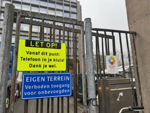 A metal fence around a school with a bright yellow sign telling pupils to leave their phones in their lockers