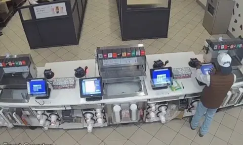 Grapevine Police Department/ Facebook CCTV shows a man tapping at a touch-screen register wearing a brown puffer vest, backward baseball cap and blue jeans.