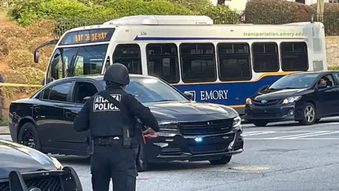 AFP via Getty Images A police officer dressed in black, with a helmet and a uniform that says "Atlanta Police" on the back, is seen standing in front of parked black sedans and a shuttle bus that says "Emory" on the side on a street.