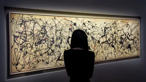 Reuters A person with long hair in silhouette is seen facing the artwork holding their mobile phone to video the painting. The artwork is long horizontally and the grey canvass appears a light tan colour under the gallery lights. It features black paint flicks and splashes all over it, together with some fewer red markings.