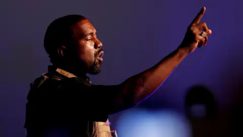 Reuters Kanye West addresses a crowd. He is a black man with a dark goatee and is wearing a military style vest. He is holding one finger up as he speaks on a stage.