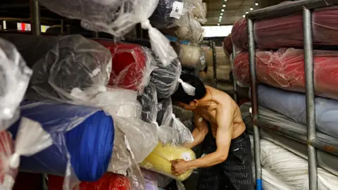 A shirtless man in black trousers wrangles large rolls of fabric wrapped in plastic, at a textile warehouse in Guangzhou