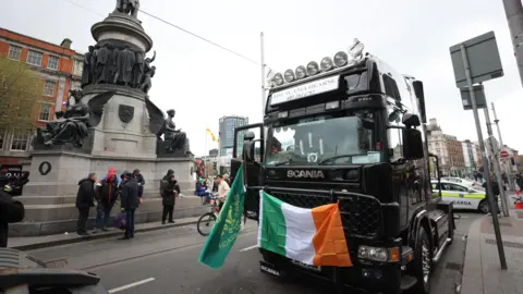 PA Media A large lorry with Irish flags attached to the front is parked in a busy street. There are men gathered around. Behind it are Garda (Irish police) cars. 