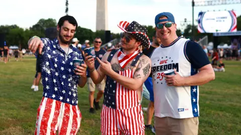 Getty Images Three men pose for the camera - the two on the left are dressed in stars and stripes, while the man on the right smiles in a UFC Freedom 250 T-shirt