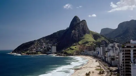 Getty Images Wide shot of Ipanema Beach in Rio De Janeiro with Morro Dois Irmaos at the end. To the left of the frame, is a row of residential buildings, to the left is the sea with waves crashing into the beach
