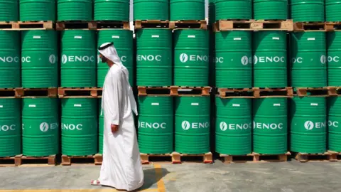 Getty Images A man wearing a long, white dishdasha walks past stacks of bright green barrels bearing