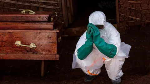 Getty Images A wooden coffin is on the left side of the image, with polished surfaces and gold-colored handles. In the foreground, a person is crouching on a dirt ground while wearing full-body protective equipment, including a white suit, a face covering, and green rubber gloves. The person’s hands are held up against the face covering, partially obscuring the face. Behind the person is a simple structure with a doorway, and the walls appear to be made of mud or brick with various wooden supports. The environment looks outdoors or semi-sheltered.