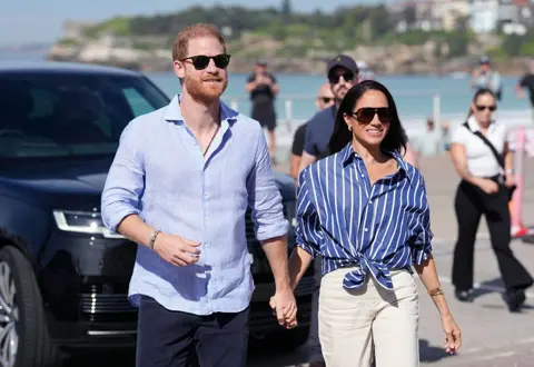 Getty Images Harry and Meghan walk together hand in hand in front of a beach in Australia.