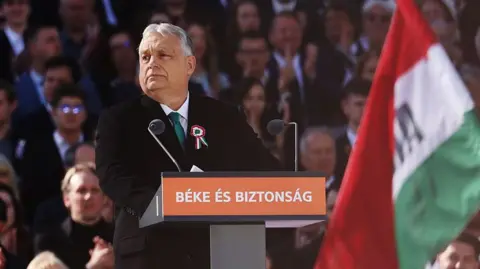 Getty Images A man in a dark suit stands at a lectern in front of a crowd and beside a flag