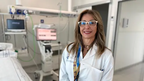 A doctor with white glasses and long brown hair smiles. She stands in a medical facility with a hospital bed next to her and two screens with several wires around them behind her.