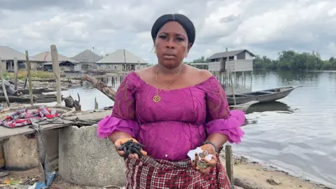 Simi Jolaoso / BBC A woman in a magenta top stands by the water and holds some dark material in her hand.