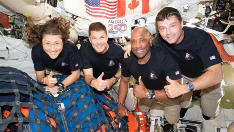 NASA Four astronauts in dark blue T‑shirts float close together inside a bright, cluttered spacecraft. Their faces are blurred, but all give thumbs‑up to the camera. Their shirts carry a matching mission patch and small US flag on the sleeve. Behind them, American and Canadian flags hang above a red‑and‑blue “America 250” logo. Around them are white padded walls, orange pipes, cables and tightly netted blue and orange cargo bags, emphasising the cramped zero‑gravity cabin.