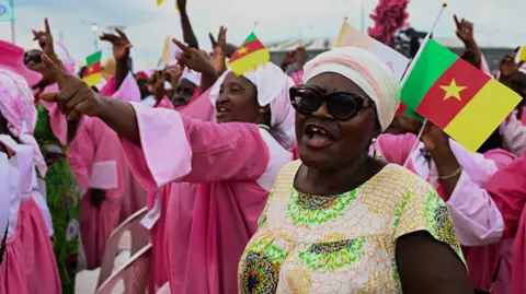 EPA A woman wearing sunglasses, a headwrap and a patterned dress in cream, green and orange, smiling and cheering. Behind her, are women wearing pink, some of whom are pointing and others are waving Cameroonian flags