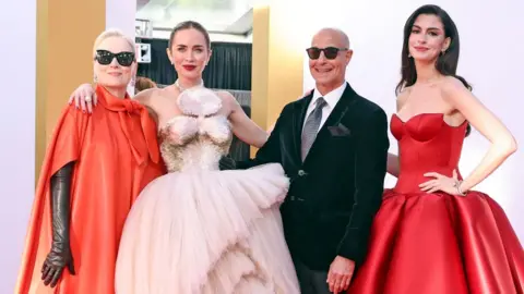 Getty Images Meryl Streep, Emily Blunt, Stanley Tucci and Anne Hathaway on the red carpet - Streep and Tucci wear dark glasses. Streep has long leather gloves and a cape-detail orange-red dress, Blunt wears a strapless pale pink gown with a layered skirt and silver detailing, Tucci is in a velvet suit jacket and Hathaway wears a strapless bright red dress with oversized skirt 