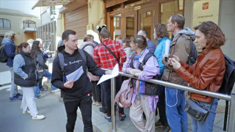 A man in a black hoodie and jeans is handing out papers to people in a queue outside an official building near the Kremlin. 