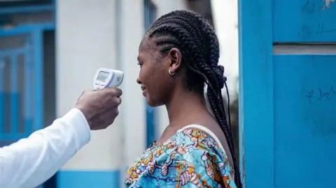 Getty Images A medic checks the temperature of a woman, using a contactless infrared thermometer. Only the arm of the medic, who is wearing white, is shown. The woman having her temperature checked has her hair in braids. She is wearing a bright blue top with a pattern and is standing next to a blue wooden door with a white and blue building in the background.