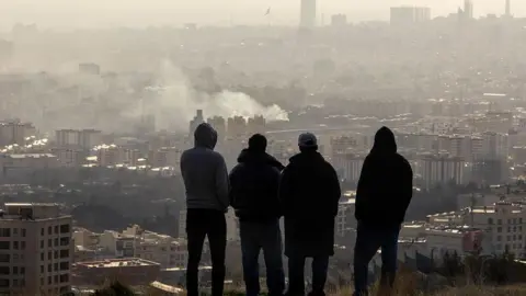 Getty Images Men watch from a hillside as a plume of smoke rises after an explosion on 2 March 2026 in Tehran, Iran.