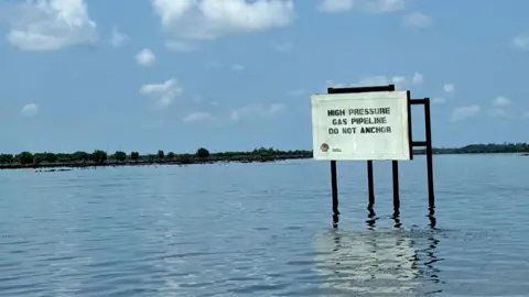 Simi Jolaoso / BBC A sign rises above the water saying: High pressure gas pipeline do not anchor.