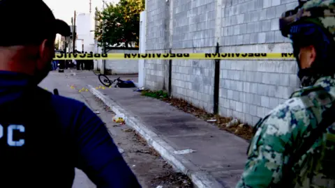 Darren Conway/BBC A soldier and another uniformed man stand behind a crime scene tape. In the distance, on the pavement, a boy is lying dead next to his bike. 