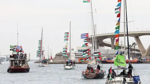 Getty Images Five small boats, each with multiple Palestinian flags, sail on the water in a Barcelona port. In the background is a large white building and the sky and water are both grey. Each boat contains several figures. 