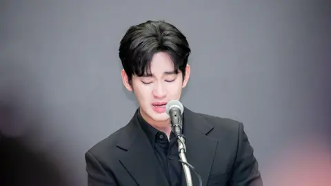 iMBC/Imazins via Getty Images A tearful Kim Soo-hyun speaking into a microphone. He is wearing a fully black outfit.