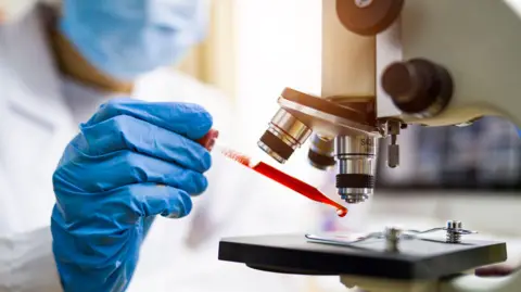 Getty Images A masked scientist wearing a blue plastic glove holds a pipette full of red liquid under a microscope. 