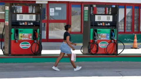 EPA/Shutterstock A woman wearing a denim skirt and a black top walks past a petrol station in Havana. There is a sign in the petrol station's window reading