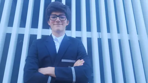 Supplied A 13-year-old boy with dark hair and glasses, wearing a school uniform of a white shirt and dark blazer, smiles at the camera.