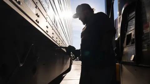 Bloomberg via Getty Images A silhouetted truck driver fills his truck against a blue sky and strong sun, with the pumps in shadow, at a gas station in Eloy, Arizona on 18 March.