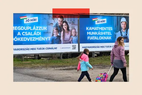 AFP via Getty Images A woman and a child walk past posters in Hungarian about pronatalist policies 