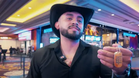 An El Temach fan wears a black open necked shirt and a black stetson hat and is holding a special chain he bought from the influencer as merchandise - it is orange with a grey geometric pattern.