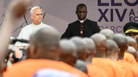 AFP via Getty Images Pope Leo in a white robe next to a cleric dressed in black looks out at a crowd of prisoners dressed in orange uniforms at Bata Prison.
