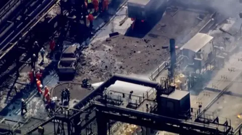 Reuters From high above, pipes, metal chimneys and walkways ring an open area with a burned out truck, mild rubble and another paneled truck. People in orange jumpsuits and white hard hats can be seen standing to one side and a fire hose blasts white jets of water.