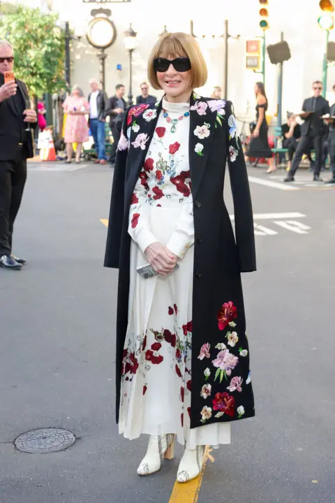 Getty Images Anna Wintour standing in a long white dress embroidered with red flowers and a long black coat, also embroidered with red and pink flowers