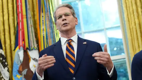 Getty Images Scott Bessent in the US Whitehouse's Oval Office. He wears a navy suit with a white shirt and striped tie, with the US official ensemble in the background.