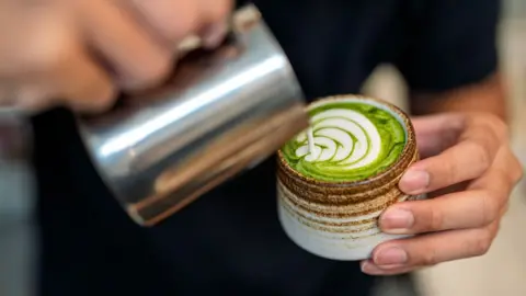 Getty Images Close-up of a barista pouring steamed milk into a ceramic cup of matcha latte, creating a pattern during preparation.