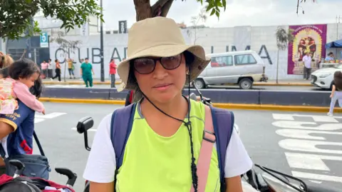 Hilda wears a yellow and white shirt and hat, with glasses and a neutral expression