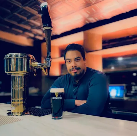 Rehan Alam Red Lion owner Rehan Alam, who has dark hair, a moustache and a beard, stands behind a bar with a well-poured pint of Guinness on it wearing a black dress shirt.