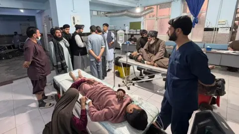 HANDOUT A young male is seen lying down on a stretcher in a hospital ward, hooked up to a drip, as a woman kneels by his side and holds his hand. A man in scrubs is seen stood next to the stretcher looking on at a group on men gathered at the end of the stretcher, while another person can be seen lying on a stretcher in the background