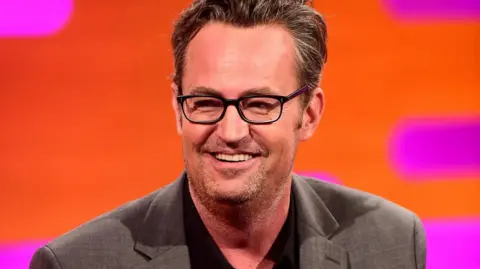PA Media Actor Matthew Perry, in dark glasses and a grey blazer, smiles against the orange and pink backdrop during filming of the Graham Norton Show in 2016.