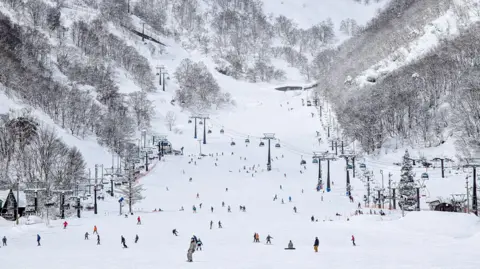 Getty Skiers dot a snowcovered mountain slope with cable cars and trees at the sides.