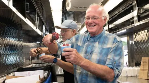 Getty Images Jerry Greenfield and Ben Cohen (right), co-founders of Ben & Jerry’s, preparing to hand out free ice cream from a van in Philadelphia. Both men are seen scooping ice cream into cups. Cohen is pictured on the right, with a smile, as he scoops ice cream. 